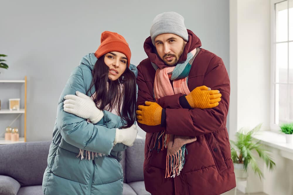 People Get Frozen Inside A Very Cold House With Electrical System And Heating Problems During Winter. Family Couple Wearing Warm Winter Clothes At Home. Husband And Wife In Coats, Hats And Gloves Looking At The Camera And Shivering