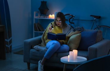 Young woman reading book with flashlight at home during blackout. She lacks a generator transfer switch to keep power running during outage