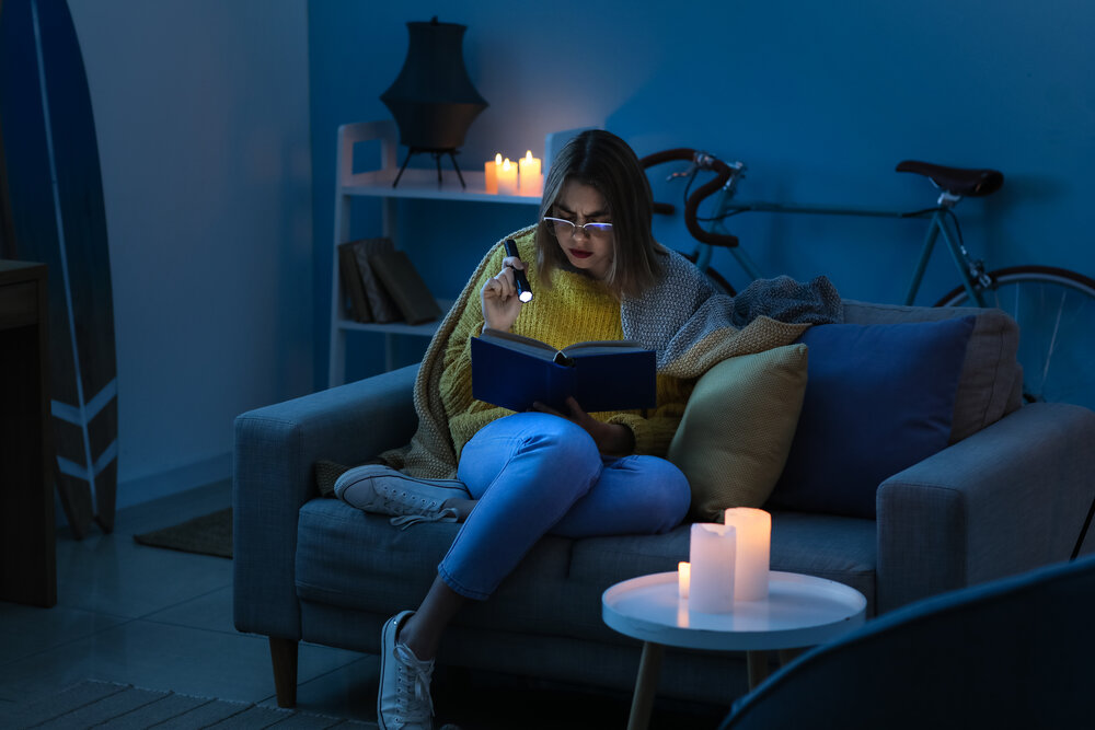 Young woman reading book with flashlight at home during blackout. She lacks a generator transfer switch to keep power running during outage