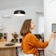 Young woman controlling home with a digital touch screen panel installed on the wall in the living room. Concept of a smart home and mobile application for managing smart devices at home. Example of 2025 remodeling trends