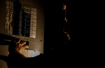 A man, visible only as a faint silhouette, is using a flashlight to investigate a home fuse box in his basement during a power outage. Circuit breaker trips when it rains.