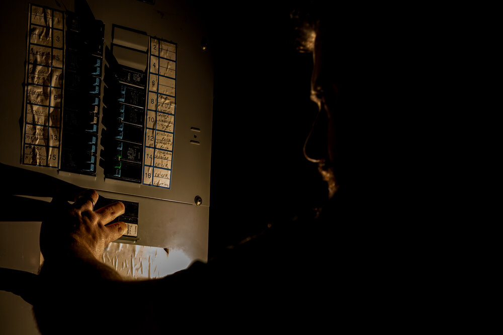 A man, visible only as a faint silhouette, is using a flashlight to investigate a home fuse box in his basement during a power outage. Circuit breaker trips when it rains.