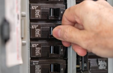 Male electrician turning off power for electrical outlet at circuit breaker box. Resetting tripped breaker in residential electricity power panel.