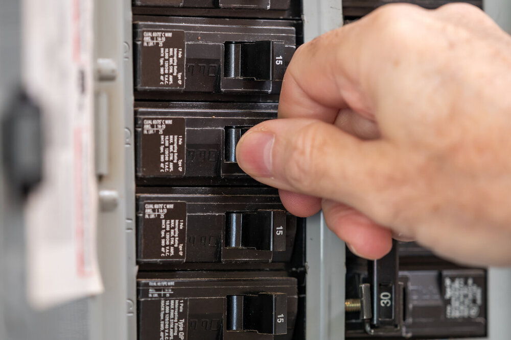 Male electrician turning off power for electrical outlet at circuit breaker box. Resetting tripped breaker in residential electricity power panel.
