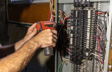 Electrician working on a circuit breaker panel is installing a ground wire using a power drill. Selective focus