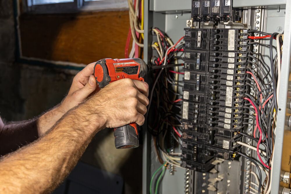 Electrician working on a circuit breaker panel is installing a ground wire using a power drill. Selective focus