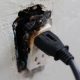 A close-up of a burnt electrical outlet with a plug still inserted, showing signs of damage from an electrical fire caused by faulty wiring.