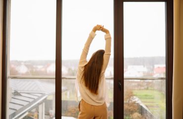 A person stretches their arms above their head while standing by a window. Outside, there are gray clouds and a view of homes and green fields.