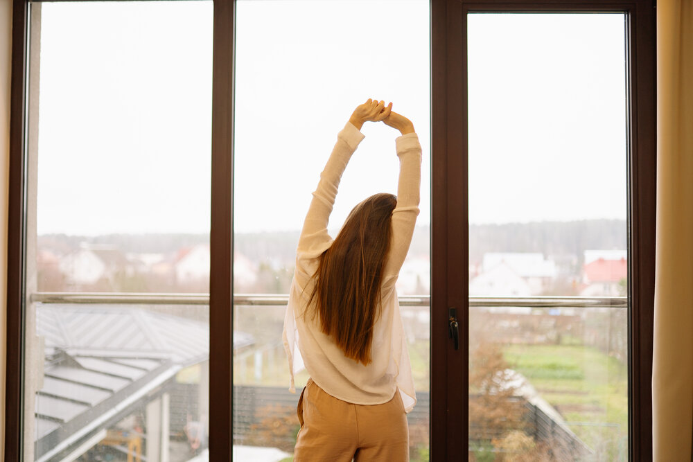 A person stretches their arms above their head while standing by a window. Outside, there are gray clouds and a view of homes and green fields.