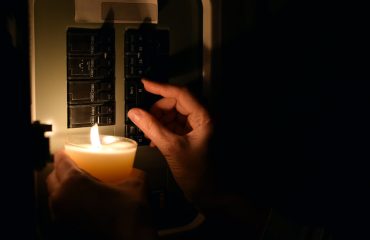 Close-up side view showing hand flipping circuit breaker switch in electrical panel while holding lit candle for illumination during blackout